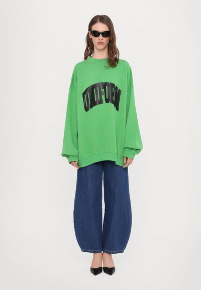 Grønn oversized genser med svart buet tekst, kombinert med vide denimbukser. Tilbehør inkluderer svarte solbriller og spisse hæler.