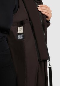 Jachetă puffer de culoare maro închis cu o textură netedă, având un buzunar interior, închidere cu fermoar și o etichetă de marcă în interior.