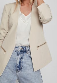 Blazer beige con textura suave, con bolsillos con cremallera, llevado sobre una camiseta blanca y combinado con vaqueros de tiro alto en azul claro.