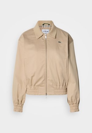Beige cropped jacket with a collared neckline, elastic cuffs, and a zipper front. Features a small green logo on the left chest.