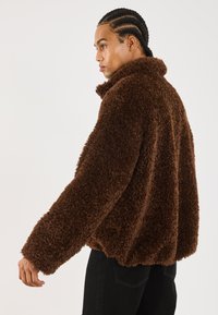 Jonge man met gevlochten haar draagt een bruine pluche fleece jas en zwarte broek, staand in profiel tegen een effen witte achtergrond.