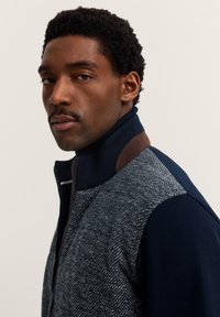 Uomo con capelli corti ricci e baffi, che indossa una giacca con zip con un motivo a spina di pesce grigio e maniche blu navy, che guarda verso la telecamera.