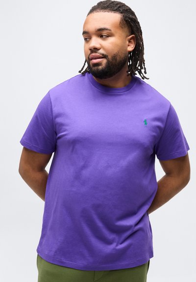 Άνδρας με ντραντλοκς που φοράει μωβ κοντομάνικο πουκάμισο και παντελόνι ελιάς, κοιτώντας ελαφρώς προς τα πλάγια με τα χέρια πίσω από την πλάτη.