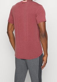 Červená melírovaná tričko s krátkým rukávem a zaobleným spodním lemem, doplněné malým logem na zadní straně, v kombinaci s šedými sportovními kalhotami se černými bočními pruhy.