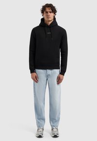 Zwarte hoodie met trekkoord en logo aan de voorkant, gecombineerd met lichtblauwe relaxed-fit jeans en grijze sneakers. Casual, comfortabele outfit.