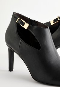 Black leather high-heeled ankle boots featuring a side cutout, gold buckle accents, and a smooth texture with a pointed toe design.