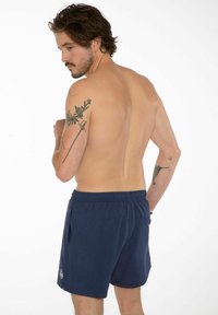 Námorníckomodré plavky s elastickým pásom, bočnými vreckami a logom, zobrazené na mužskom modeli s odhalenou pokožkou z pohľadu zozadu.