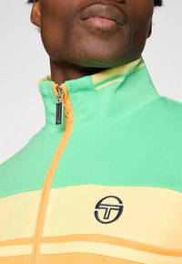 Grønn, gul og oransje zip-up treningsjakke. Har høy krage, stripete mønster og logo på brystet. Glatt tekstur.
