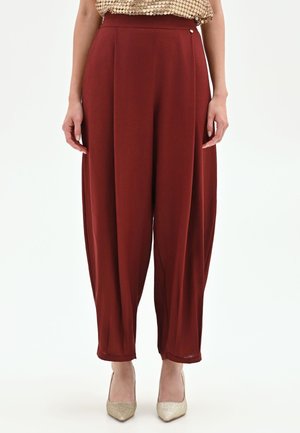 Donna che indossa pantaloni a vita alta a gamba larga color ruggine e tacchi dorati a punta, in piedi davanti a uno sfondo bianco.