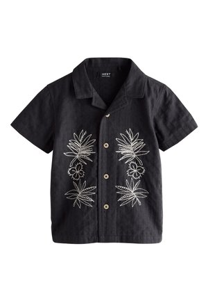Ung dreng i en sort skjorte med hvide motiver og beige shorts står på jord nær et appelsintræ og kigger til siden.
