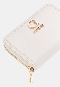 Portafoglio in ecopelle bianca con zip dorata, caratterizzato da cuciture impresse e una placca con il logo dorato che recita "GUESS". Forma compatta e rettangolare.