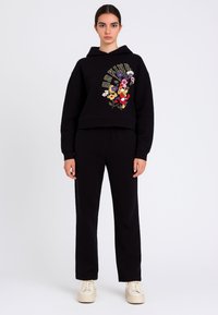 Felpa corta nera con ricamo floreale e scritte dorate, abbinata a pantaloni neri e scarpe color crema. Tessuto dalla texture liscia.