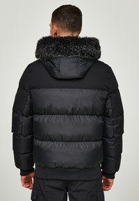 Schwarze Steppjacke mit Kapuze, verfügt über einen Kunstpelzkragen, gerippte Bündchen und gesteppte Einsätze zur Isolierung. Strukturierte Oberfläche, leichtes Design.