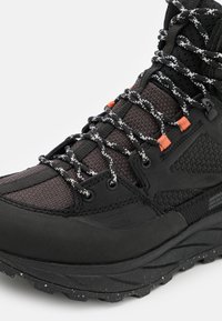 Stivale da hiking nero con tomaia testurizzata, dettagli arancioni, lacci riflettenti e una suola robusta con un motivo maculato.