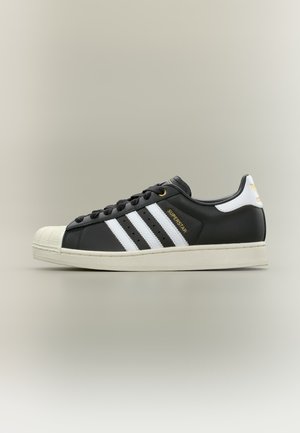 Scarpa Adidas Superstar nera con strisce bianche, punta in gomma, suola bianca, scritta dorata "Superstar" e logo sul tallone.