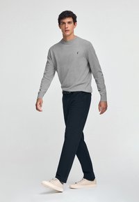 Maglione grigio a maglia, pantaloni navy e sneakers chiari. Logo distintivo sul lato sinistro del maglione. Modello che cammina con le mani rilassate.
