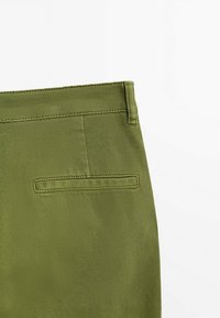 Gros plan sur un tissu vert olive avec une poche passepoilée simple et une boucle de ceinture, mettant en valeur les coutures détaillées et la texture.