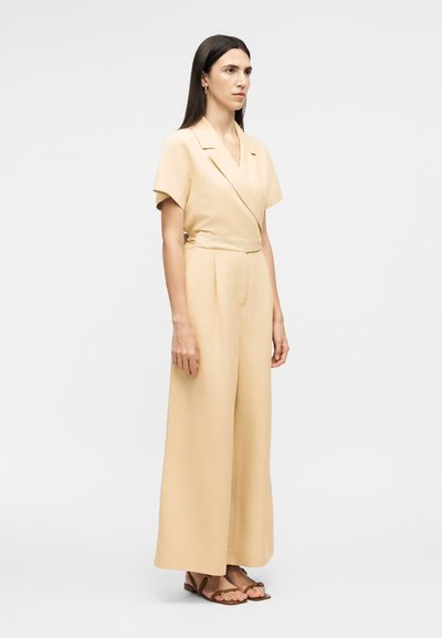 Γυναίκα με μακριά σκούρα μαλλιά, φορώντας μπεζ ολόσωμη φόρμα με κοντά μανίκια, γιακά και παντελόνι με φαρδιά πόδια, στέκεται αντικρίζοντας προς τα εμπρός σε ουδέτερο φόντο.