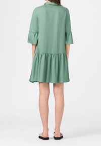 Zielona sukienka do kolan, uszyta z gładkiego materiału, z falbaną na brzegu i szerokimi rękawami w kształcie dzwonu, z kołnierzykiem z tyłu.