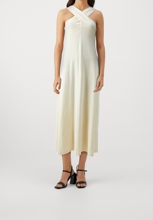 Kvinde iført en ærmeløs midi-kjole i cremefarve med krydsede stropper ved halsudskæringen og sorte højhælede sandaler.