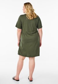 Robe t-shirt verte, longueur genoux, avec des manches courtes, coupe décontractée, taille cintrée et texture douce. Portée avec des sandales noires à enfiler.