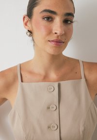 Bež obleka brez rokavov s kvadratnim izrezom, s tremi gumbi spredaj, gladko teksturo in minimalističnim dizajnom.