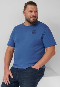 Niebieska bawełniana koszulka z krótkim rękawem, okrągłym dekoltem i czarnym logo na lewej piersi. Połączona z ciemnoniebieskimi dżinsami.