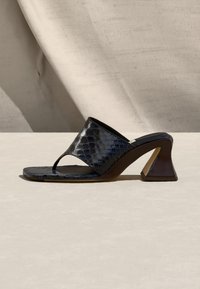 Μαύρο πέδιλο με ψηλό τακούνι, υφασμένη φαρδιά λουρίδα και δάχτυλο μπροστά, σε μπεζ φόντο με απαλά σκιάσεις.