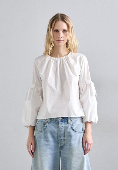 Chemisier blanc à manches longues avec un col froncé et des manches bouffantes, agrémenté de nœuds. Porté avec un pantalon en denim bleu clair, ample.