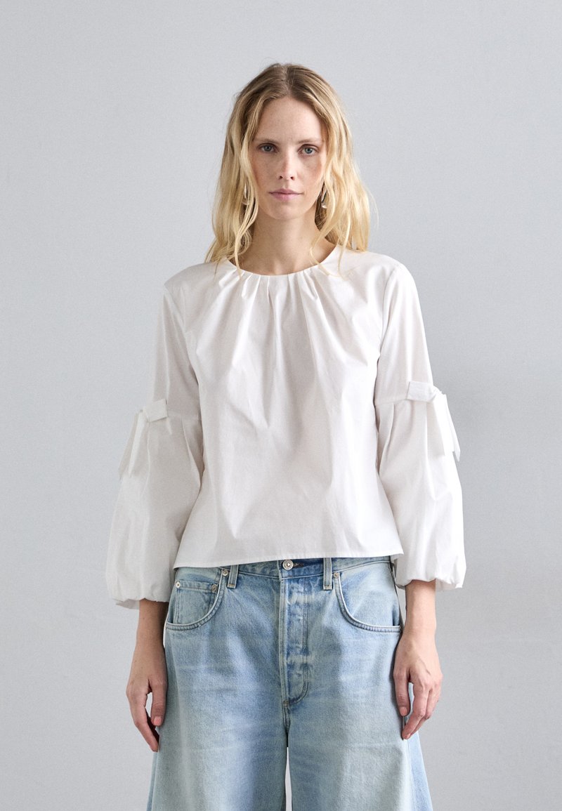 Fehér, hosszú ujjú blúz, összegyűrt nyakkal és puffos ujjakkal, masni díszítéssel. Világoskék, laza szabású farmernadrággal viselve.