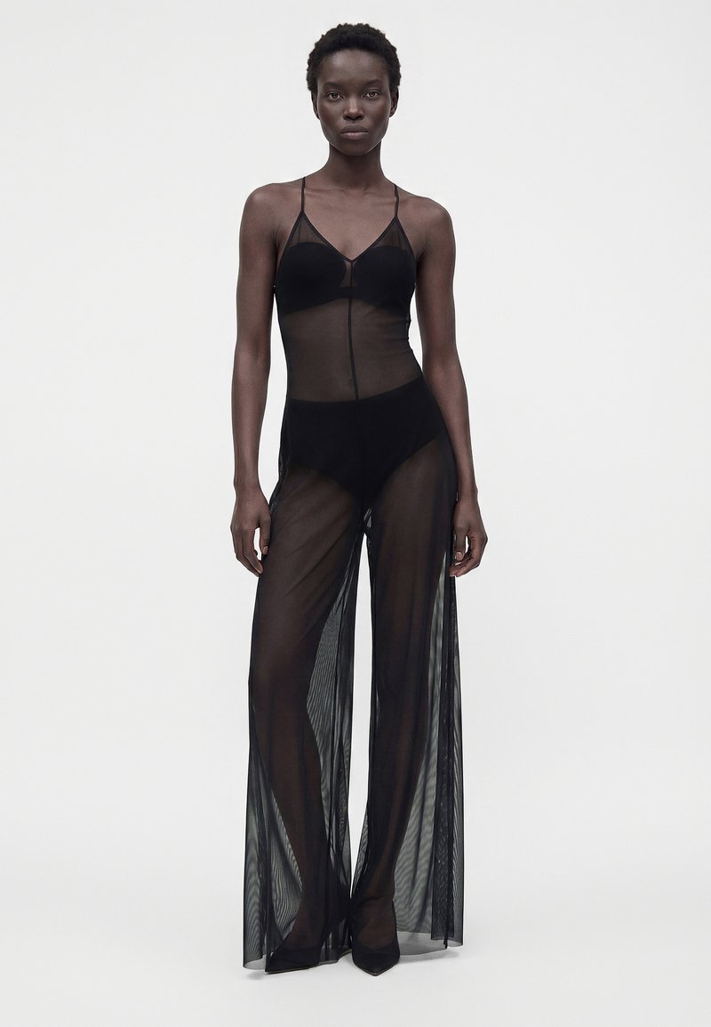 Femme noire portant une combinaison noire transparente avec des bretelles spaghetti et un pantalon large, debout devant un fond clair uni.