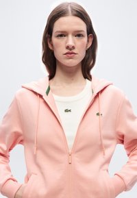 Νεαρή γυναίκα με μακριά μαλλιά, φορώντας ροζ φούτερ με φερμουάρ πάνω από ένα λευκό πουκάμισο με πράσινα λογότυπα κροκοδείλων, στέκεται με τα χέρια στις τσέπες.