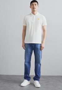 Cămașă polo albă cu un emblema galbenă, guler ribbat cu accente deschise, purtată cu blugi denim albaștri și adidași albi.