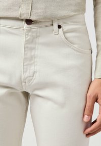 Jeans in denim beige con una vestibilità slim, dotati di tasche frontali, cuciture sottili e un'etichetta marrone in vita. Texture liscia.