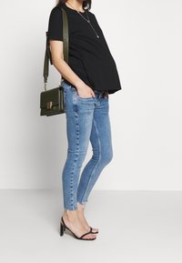 Mustat lyhyet hihatoput raskaana, siniset kapeat farkut, joissa on revityt helmat, mustat remmikorot ja tummanvihreä olkalaukku kultaisilla yksityiskohdilla.