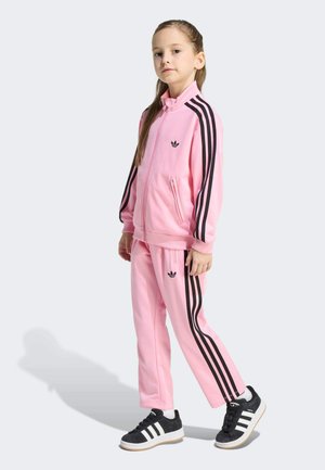 Young girl standing wearing a pink Adidas tracksuit with black stripes and black and white Adidas sneakers on a light background.