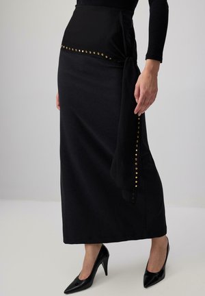 Gonna maxi nera con design a wrap e finitura in satin, decorata con borchie dorate lungo la vita e il lato, abbinata a tacchi neri.