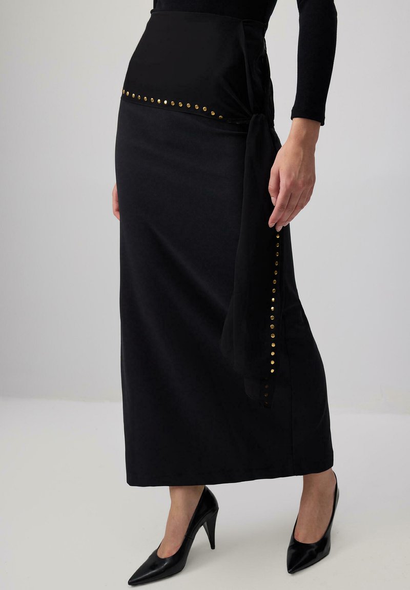 Gonna maxi nera con design a wrap e finitura in satin, decorata con borchie dorate lungo la vita e il lato, abbinata a tacchi neri.