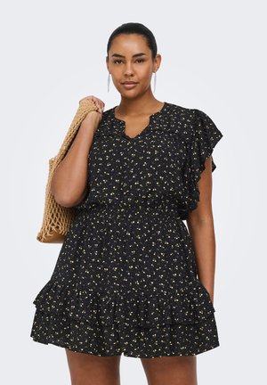 Femme portant une robe noire à fleurs avec des volants, tenant un sac fourre-tout tissé beige sur son épaule, faisant face à l'avant avec une expression neutre.