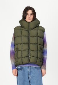 Vesta din puf de culoare verde măslinie, cu guler înalt, textură matlasată, închidere frontală cu fermoar și buzunare laterale, purtată peste un pulover tricotat multicolor.