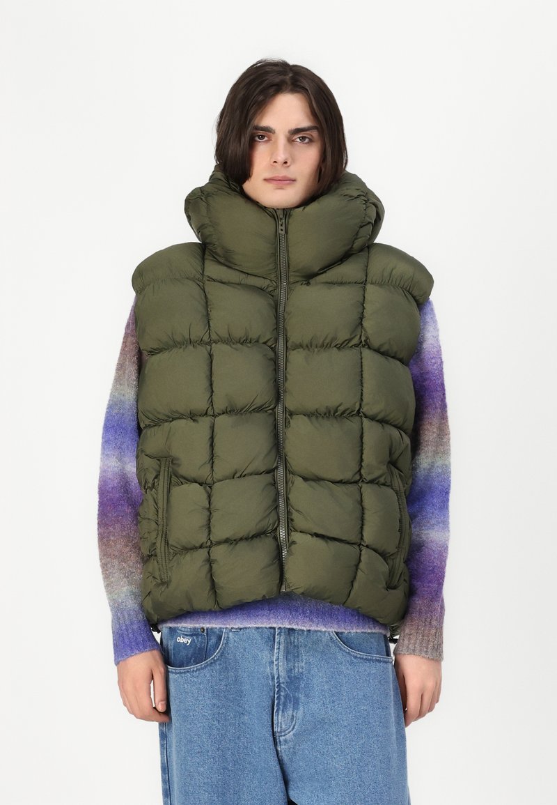 Vesta din puf de culoare verde măslinie, cu guler înalt, textură matlasată, închidere frontală cu fermoar și buzunare laterale, purtată peste un pulover tricotat multicolor.