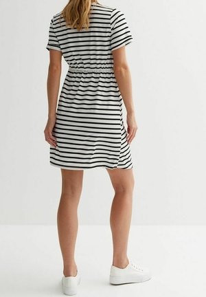 Femme debout de dos, portant une robe courte à manches courtes, rayée noir et blanc atteignant les genoux, avec des baskets blanches sur un fond blanc.