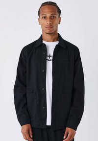 Zwarte button-up jas met twee voorkeurszakken, gemaakt van een gestructureerde stof, gedragen over een witte T-shirt met een grafisch ontwerp.