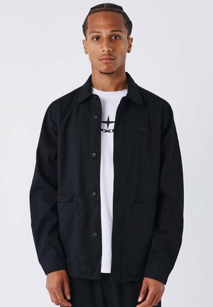 Chaqueta negra de botones con dos bolsillos frontales, hecha de un tejido texturizado, llevada sobre una camiseta blanca con un diseño gráfico.