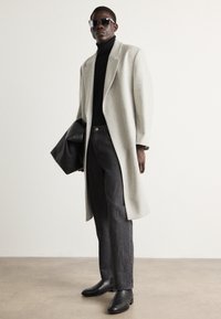 Manteau en laine gris clair, col roulé noir, jean en denim foncé, bottes en cuir noires et lunettes de soleil. Couleur unie, design moderne, coupe structurée.