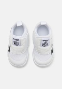 Bijele Reebok tenisice s glatkom teksturom, s čičak trakama i crnim detaljima. Lagani dizajn s zaobljenim vrhom i podstavljenim unutrašnjim dijelom.