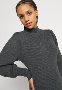 Suéter de canalé gris oscuro con cuello alto, mangas abullonadas y diseño ajustado. La textura es suave con un brillo sutil.