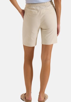 Person trägt beige Shorts mit elastischem Bund und braune Slipper, steht mit einer Hand an der Hüfte, Rückansicht.