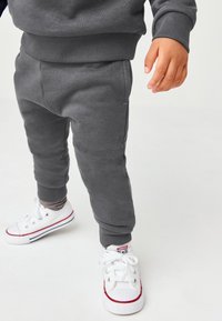 Graue Baumwoll-Sweatpants mit gerippten Bündchen, kombiniert mit weißen Turnschuhen mit roten Streifen und einem Logo, wobei eine gestreifte Socke darunter sichtbar ist.