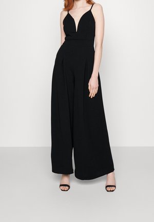 Woman wearing a sleeveless black jumpsuit with wide-leg pants and black open-toe heels against a plain white background.